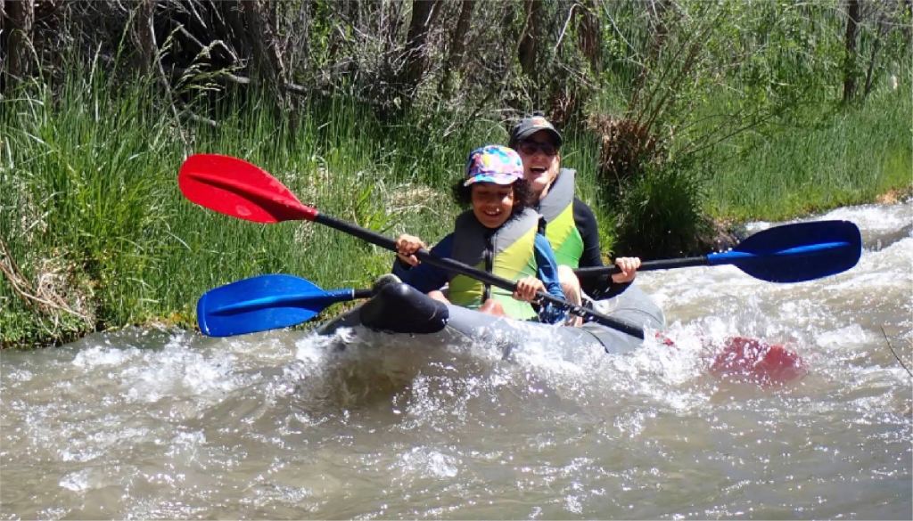 Two people kayaking on a narrow, fast-flowing river with red and blue paddles.