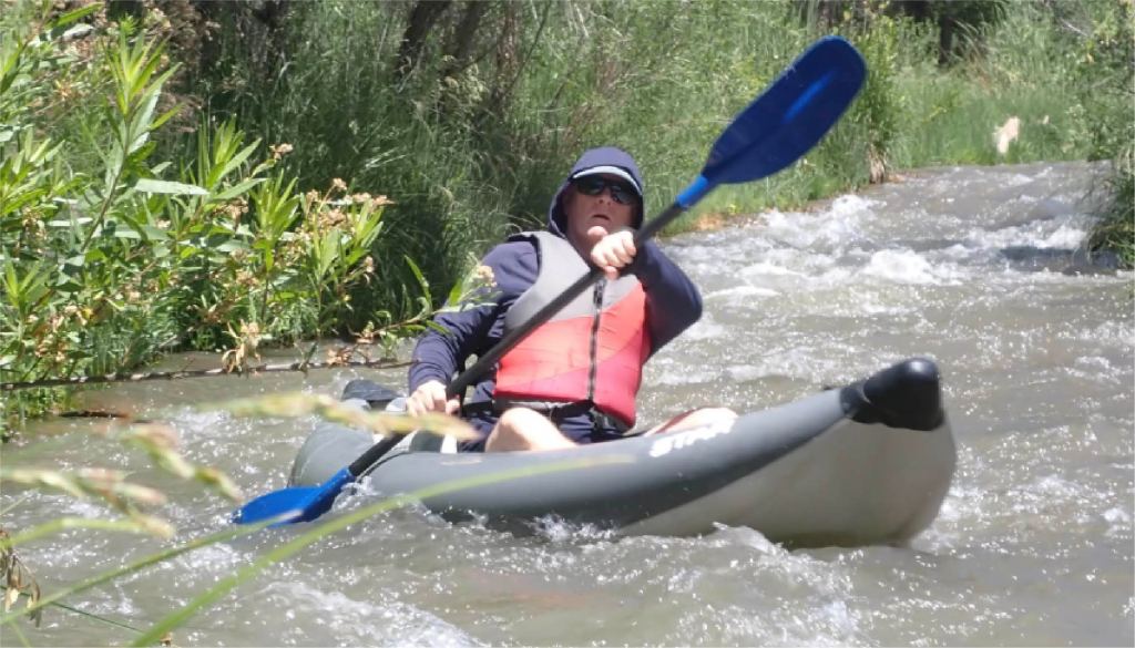 Person kayaking down a narrow, rocky stream with greenery on the banks.