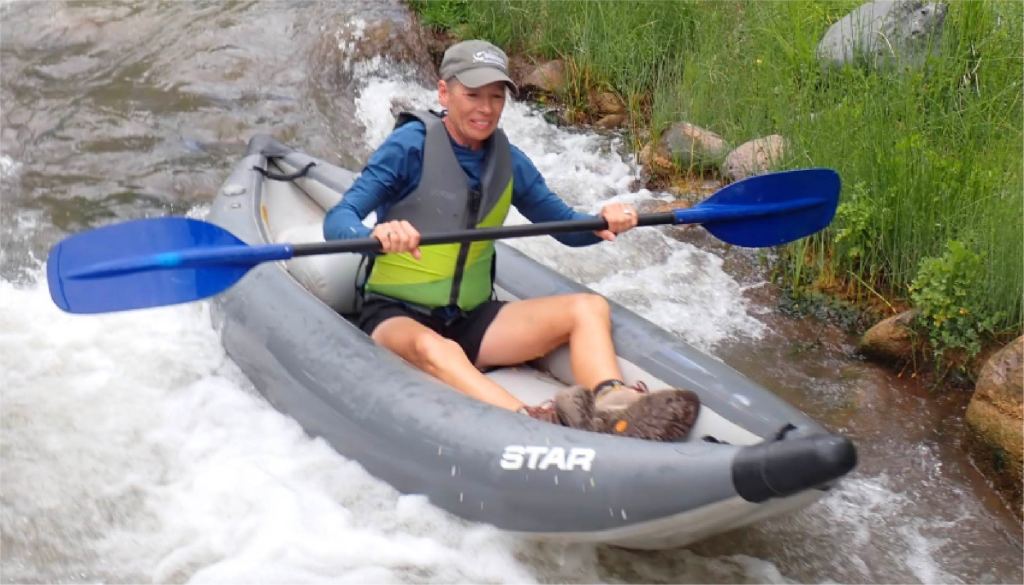 Person kayaking on a river, wearing a life jacket and cap, holding blue paddles.