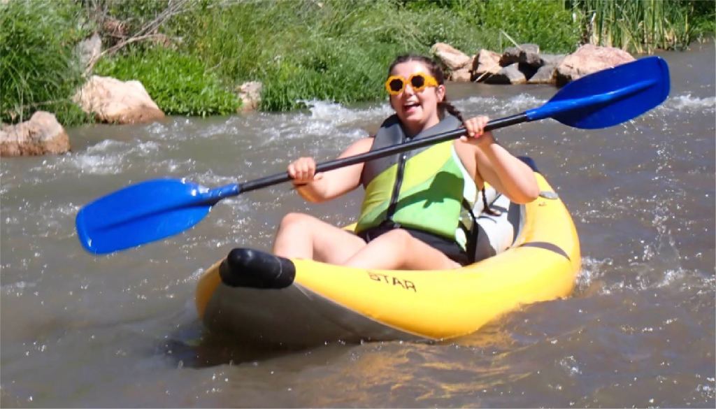 Person kayaking on a river, wearing a life jacket and sunglasses.