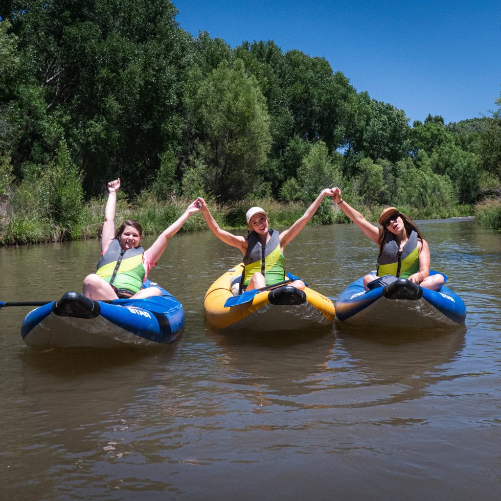 Three people in life vests on inflatable kayaks holding hands on a river.