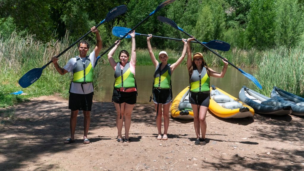Four people in life vests hold paddles above their heads, standing near inflatable kayaks by a river.