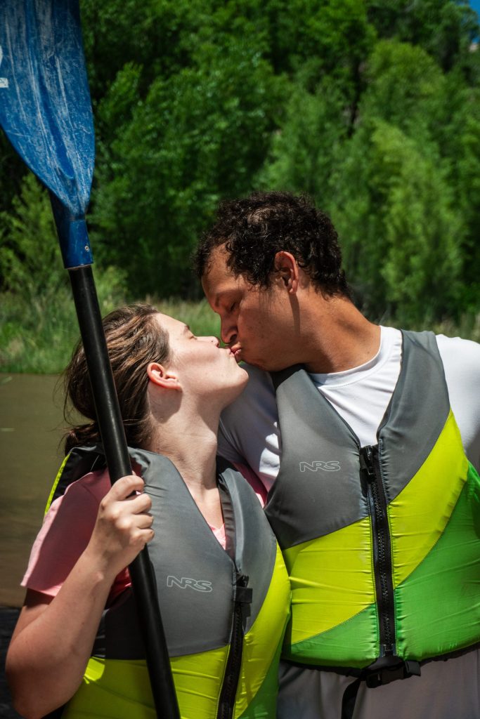 Couple in life vests kissing by river with a paddle.