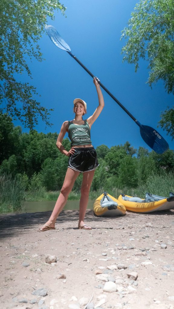 Person holding kayak paddle standing on a riverside with two yellow kayaks.