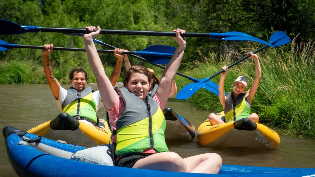 Three people kayaking on a river, holding paddles up, wearing life vests.