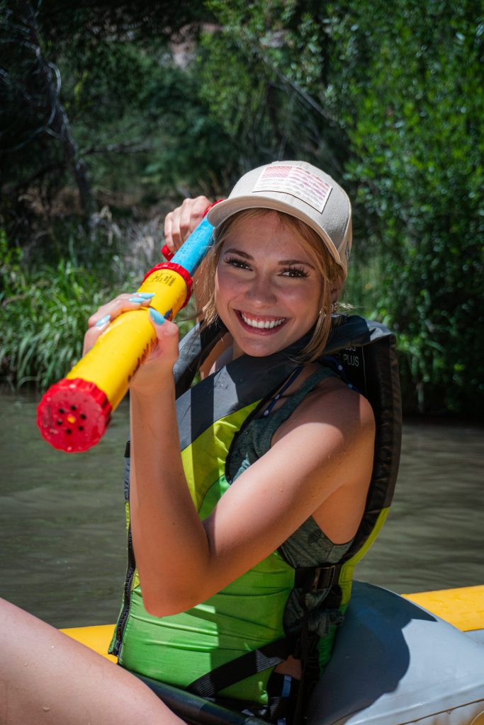 Person in life vest and cap smiling, holding a colorful water blaster in a lush outdoor setting.