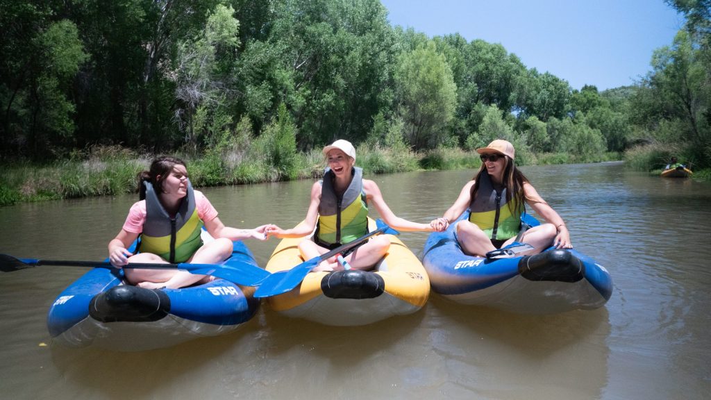Three people in kayaks holding hands on a calm river.