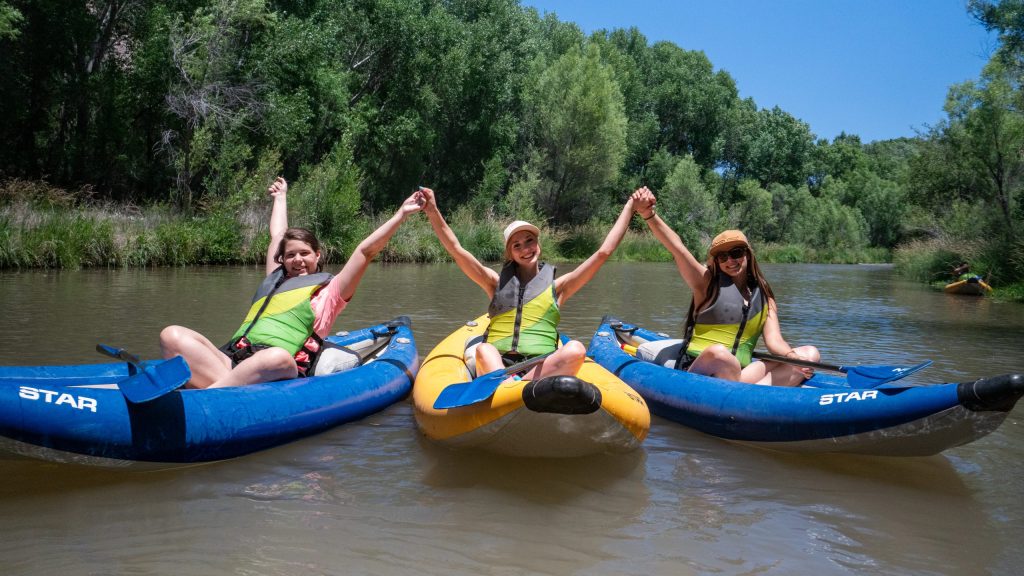 Three people in kayaks holding hands on a calm river, surrounded by trees.