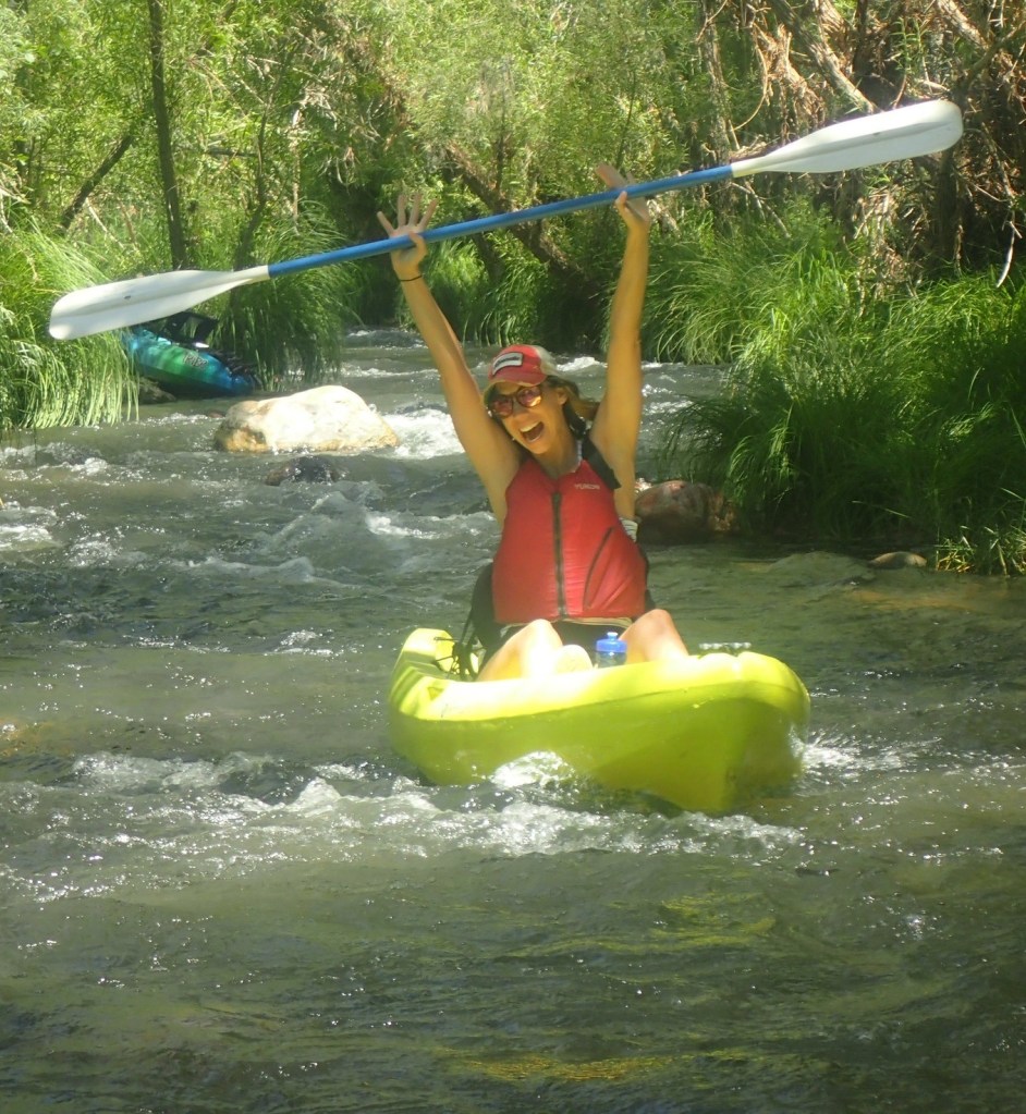 Person in a kayak holding a paddle aloft, navigating a narrow stream.