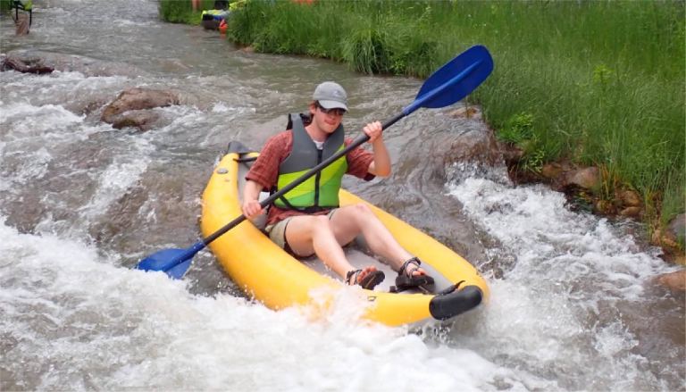 Person kayaking through a rocky stream in a yellow kayak, wearing a life vest and cap.