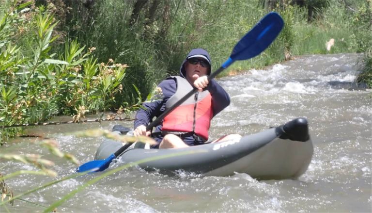Person kayaking down a narrow, rocky stream with greenery on the banks.