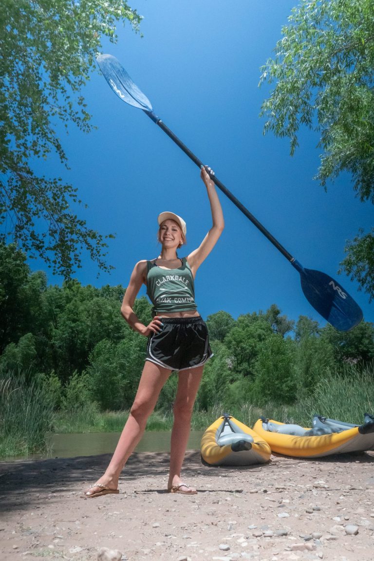 Person holding kayak paddle standing on a riverside with two yellow kayaks.